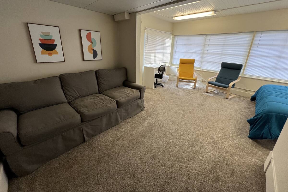 Una habitación con un sofá marrón, una silla amarilla y una cama azul. Hay dos obras de arte enmarcadas en la pared.