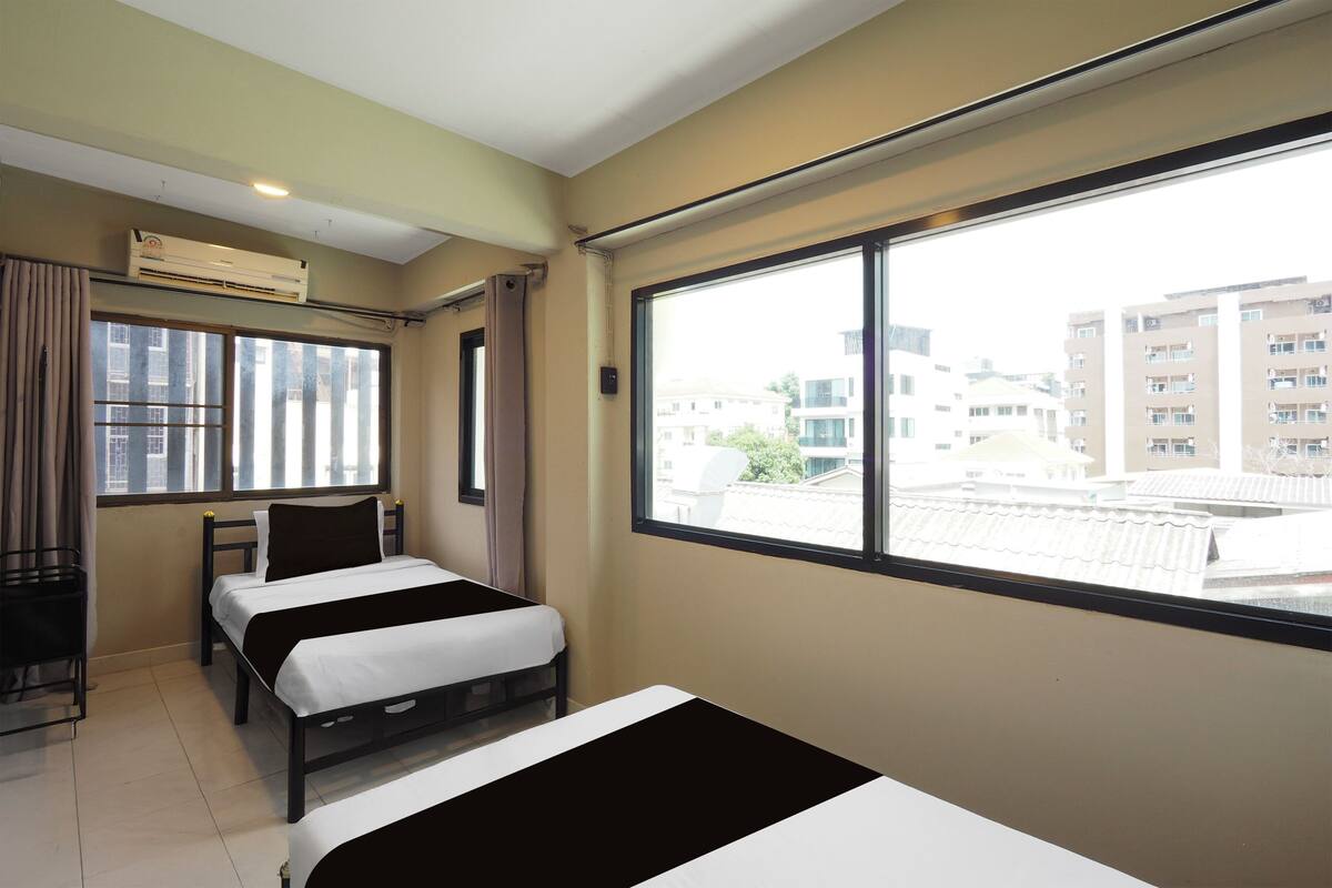 A hotel room with two beds, a window with a view of buildings, an air conditioning unit, and a desk with a chair.