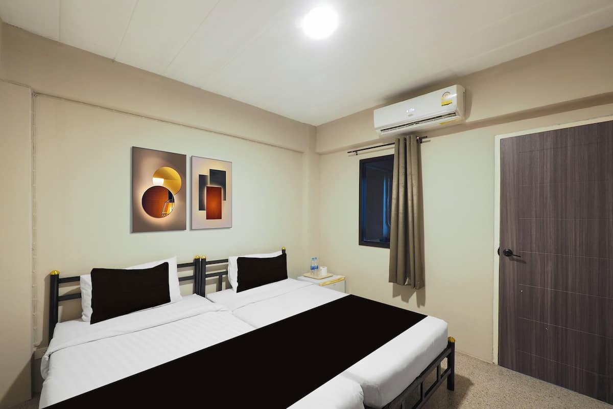 A hotel room with a double bed, a bedside table with a bottle, an air conditioning unit, a door, and two wall-mounted artworks.