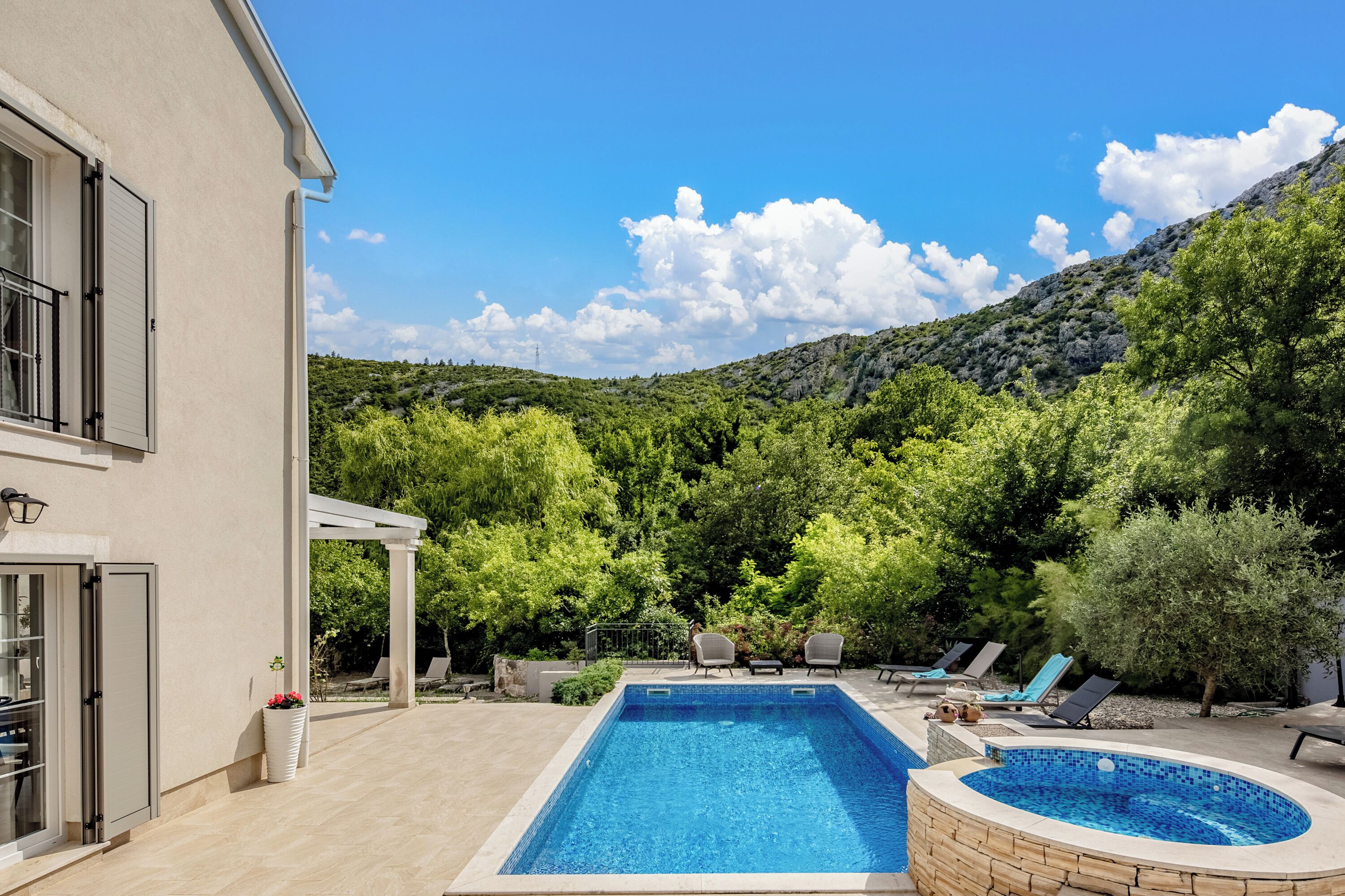 A rectangular swimming pool with a stone edge, surrounded by lounge chairs and a pergola, set against a backdrop of lush greenery and a mountain.