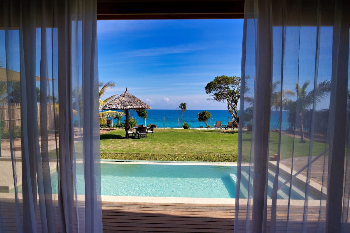 Blick von einem Zimmer mit Pool auf einen Strand mit einer Reetdachsonnenliege und Stühlen.