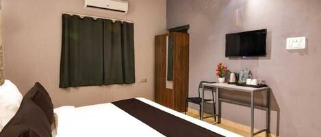 A hotel room with a bed, an air conditioning unit, a TV, a desk with a chair, and a wardrobe.