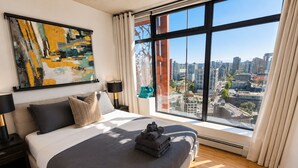 Ein modernes Hotelzimmer mit einem großen Bett, Blick auf die Stadt, einem Gemälde an der Wand und einem Nachttisch mit einer Lampe.