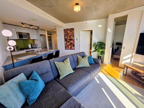 Una sala de estar moderna con un sofá seccional grisáceo, una cocina al fondo y una zona de comedor con una mesa de madera.