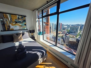 Una habitación de hotel moderna con una cama grande, vistas a la ciudad, un cuadro en la pared y cortinas en la ventana.