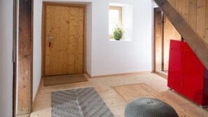 Een gang met een houten deur, een klein raam met een plant, een rode kast en een grijs vloerkleed.