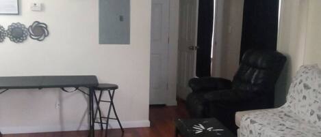 A living room with a sofa, a chair, a small table, and a bench. There is a door leading to another room.