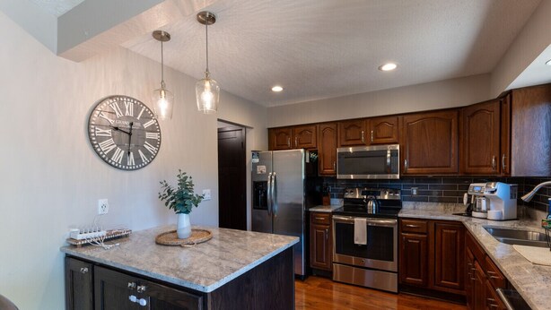 A modern kitchen with a central island, stainless steel appliances, wooden cabinets, pendant lighting, and a large wall clock. - Hot Tub | Built in Fire Pit | Fenced Yard for Kids (Fargo)