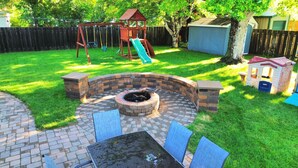 A backyard with a brick fire pit, outdoor seating, a swing set, and a slide. - Hot Tub | Built in Fire Pit | Fenced Yard for Kids (Fargo)