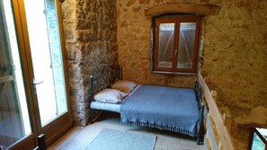 A small, rustic bedroom with a stone wall, a wooden bed frame, a single window with wooden shutters, and a door. - Tournemire Vine House (TOURNEMIRE)
