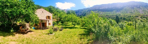 Une maison en pierre avec un porche en bois, entourée d’une végétation luxuriante et avec des montagnes en arrière-plan.