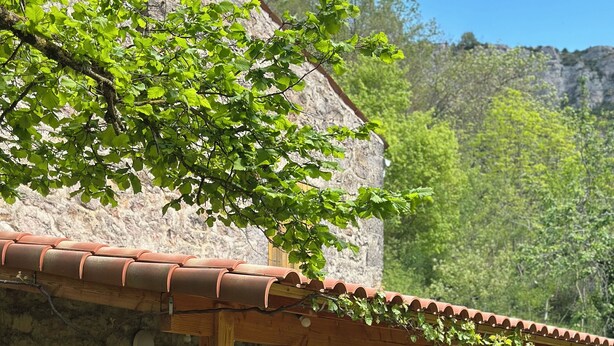A rustic stone building with a wooden pergola, picnic tables, and a stone wall.
