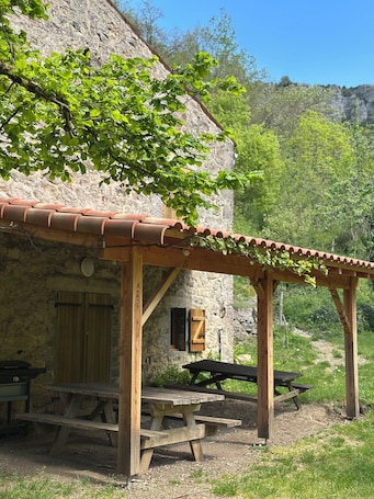 A rustic stone building with a wooden pergola, picnic tables, and a stone wall.. Tournemire Vine House