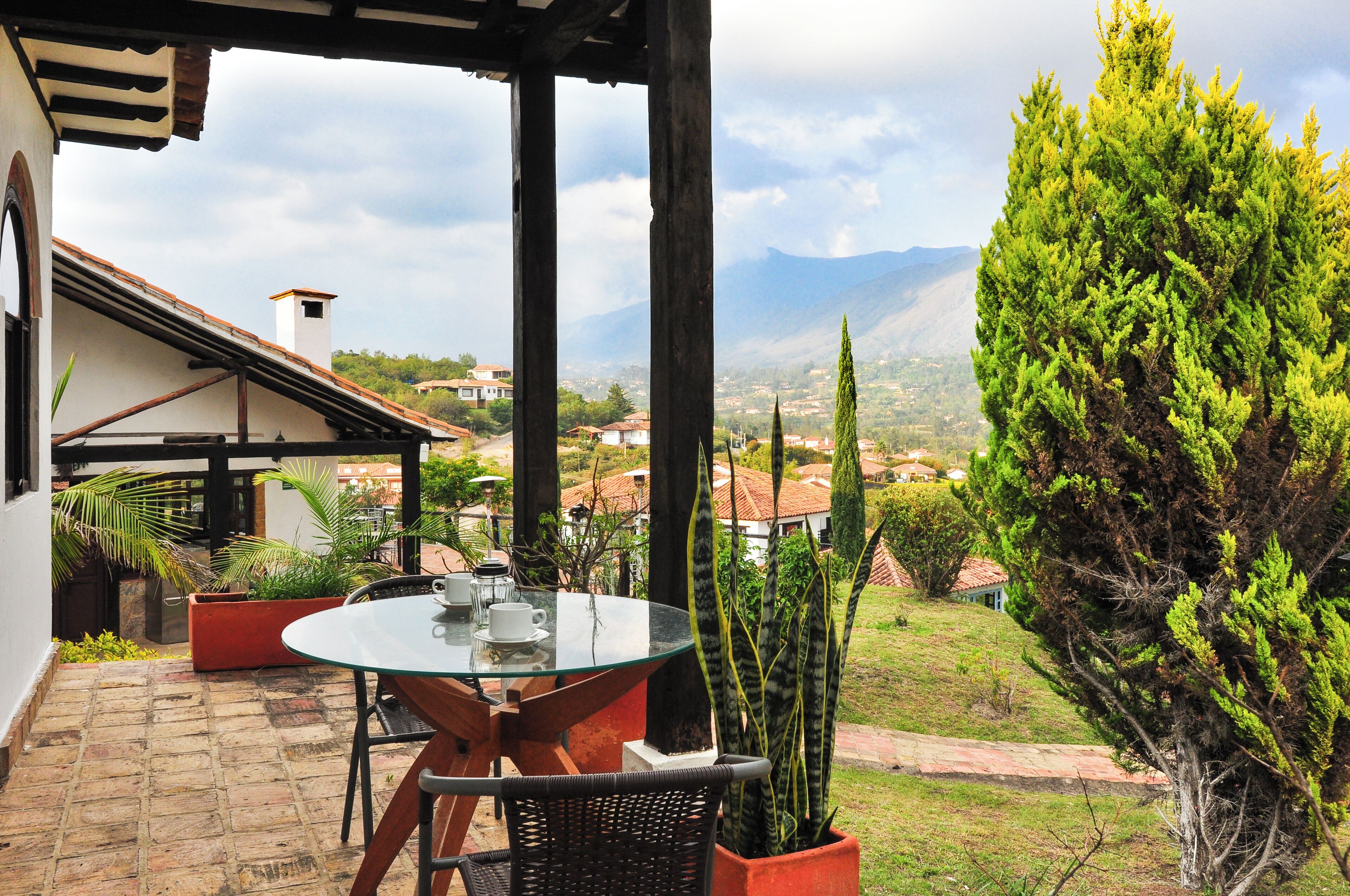 Un patio con una mesa y sillas de vidrio, rodeado de plantas en macetas y vistas a un pueblo con montañas al fondo.