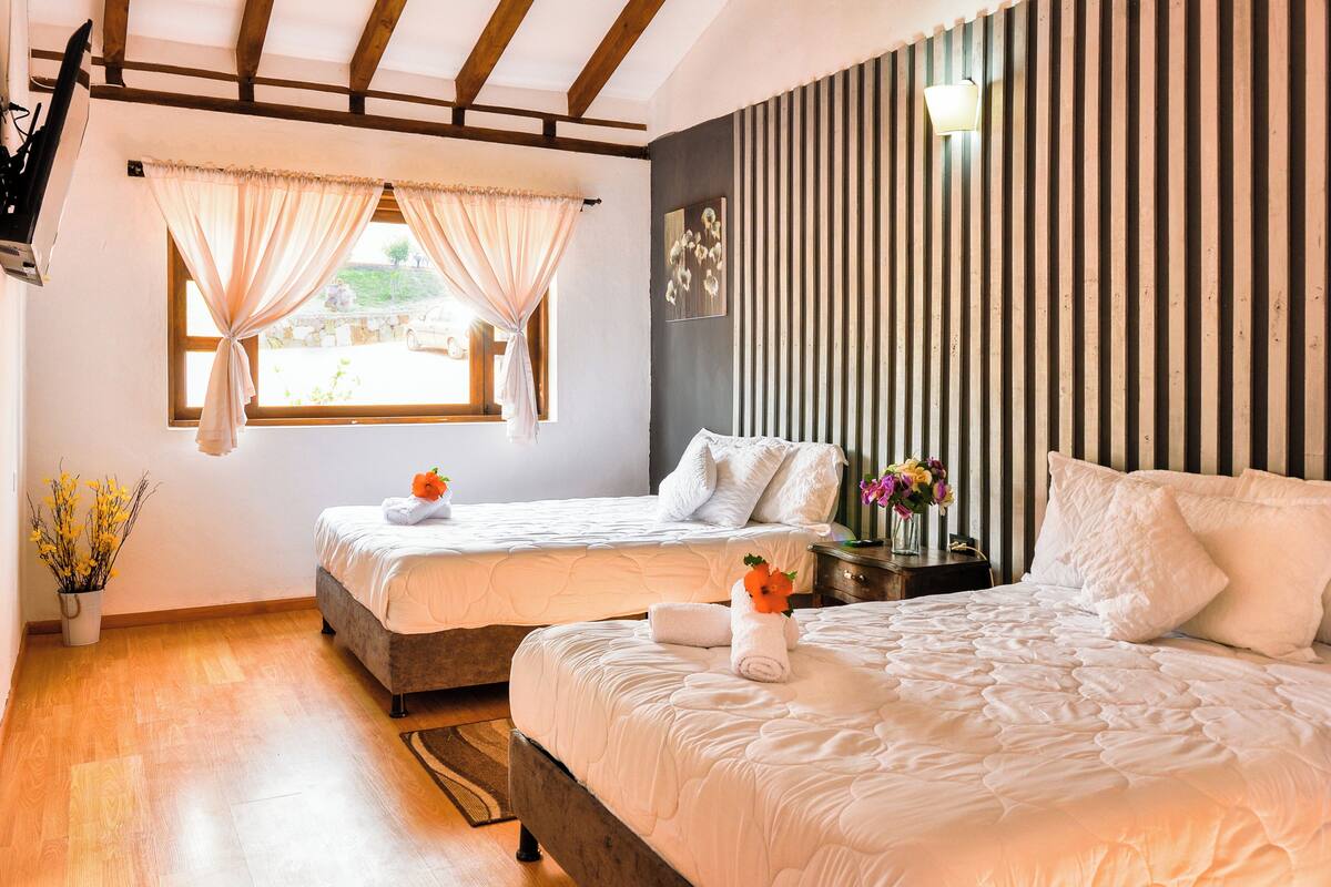 Une chambre d’hôtel avec deux lits, un plafond en bois, une fenêtre avec des rideaux, une table de chevet avec une lampe et un vase de fleurs.