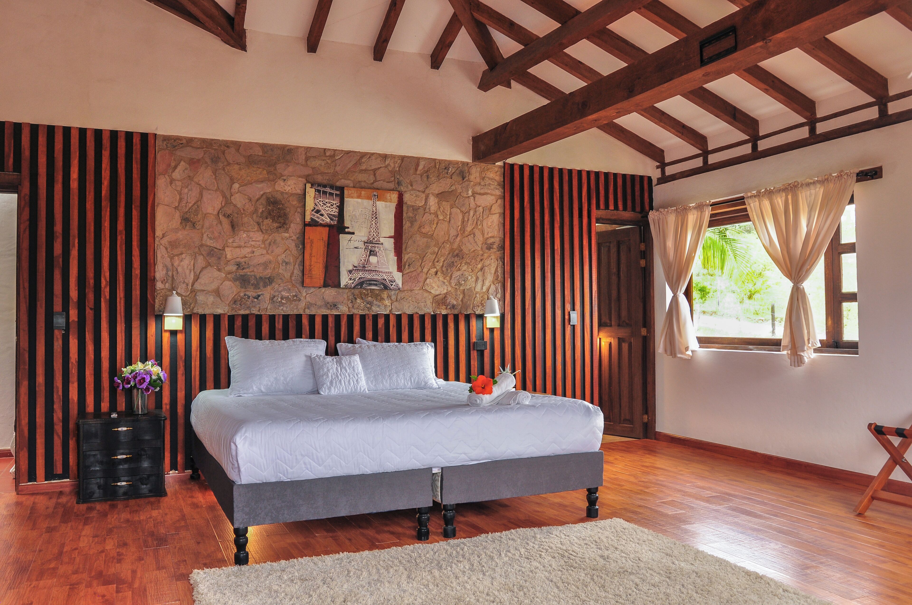 A bedroom with a stone accent wall, a bed with white bedding, a nightstand with a lamp, and a window with curtains.