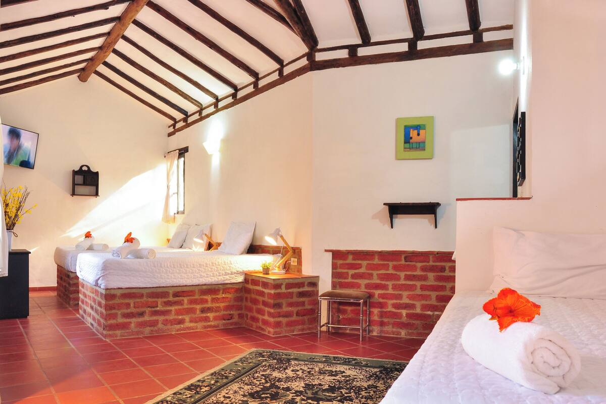 Une pièce avec un mur en briques, un lit avec une serviette roulée, un petit tabouret, une table de chevet avec une lampe et une fenêtre avec des rideaux.