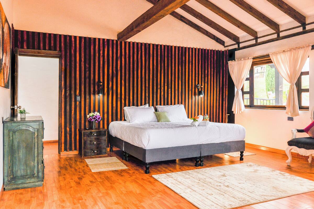 Une chambre à coucher avec un lit, un plancher en bois, une commode, un miroir et une fenêtre avec des rideaux.
