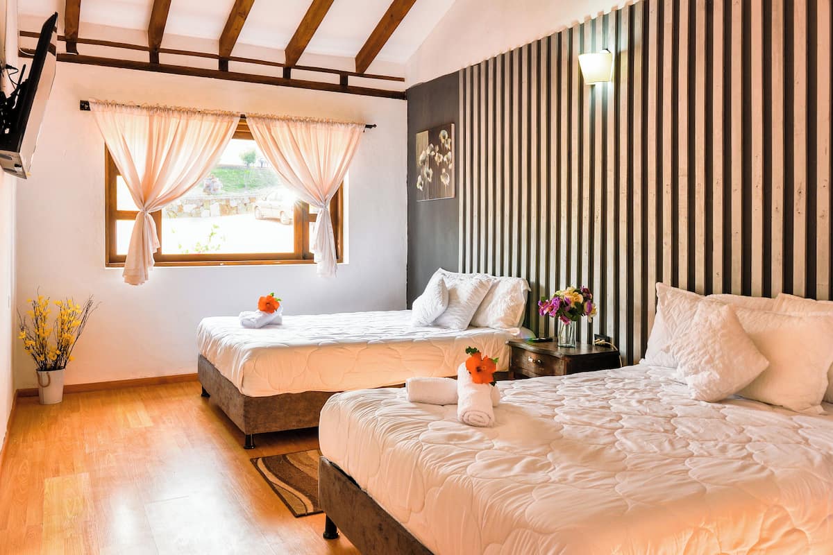 Une chambre d’hôtel avec deux lits, un plafond en bois, une fenêtre avec des rideaux, une table de chevet avec une lampe et un vase de fleurs.