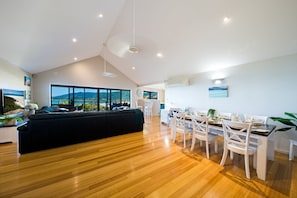 Un vaste espace de vie comprenant un grand canapé noir, une table à manger avec des chaises blanches et une vue sur les montagnes, visible à travers de grandes fenêtres.