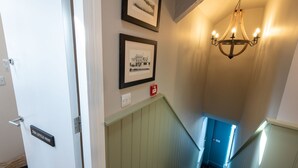 A staircase with a blue door at the top, a chandelier, and two framed pictures on the wall. - The Little Nest : Charming 2-Bed Hideaway by Sea in Rhoscolyn | Managed by Anglesey Holiday Lettings (Rhoscolyn)