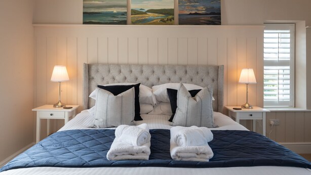 A neatly made bed with a quilted blue and white duvet, two bedside lamps, a tufted headboard, and three landscape paintings above the bed. - The Little Nest : Charming 2-Bed Hideaway by Sea in Rhoscolyn | Managed by Anglesey Holiday Lettings (Rhoscolyn)