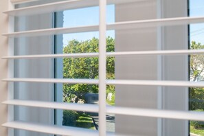 A window with horizontal blinds, through which a tree-lined street is visible.