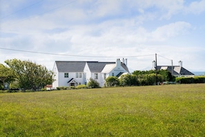 A white building with a gabled roof and chimneys, surrounded by a grassy field and trees.