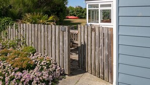 Ein hölzernes Tor führt zu einem Haus mit einem Fenster, umgeben von einem Garten mit Blumen und Bäumen.