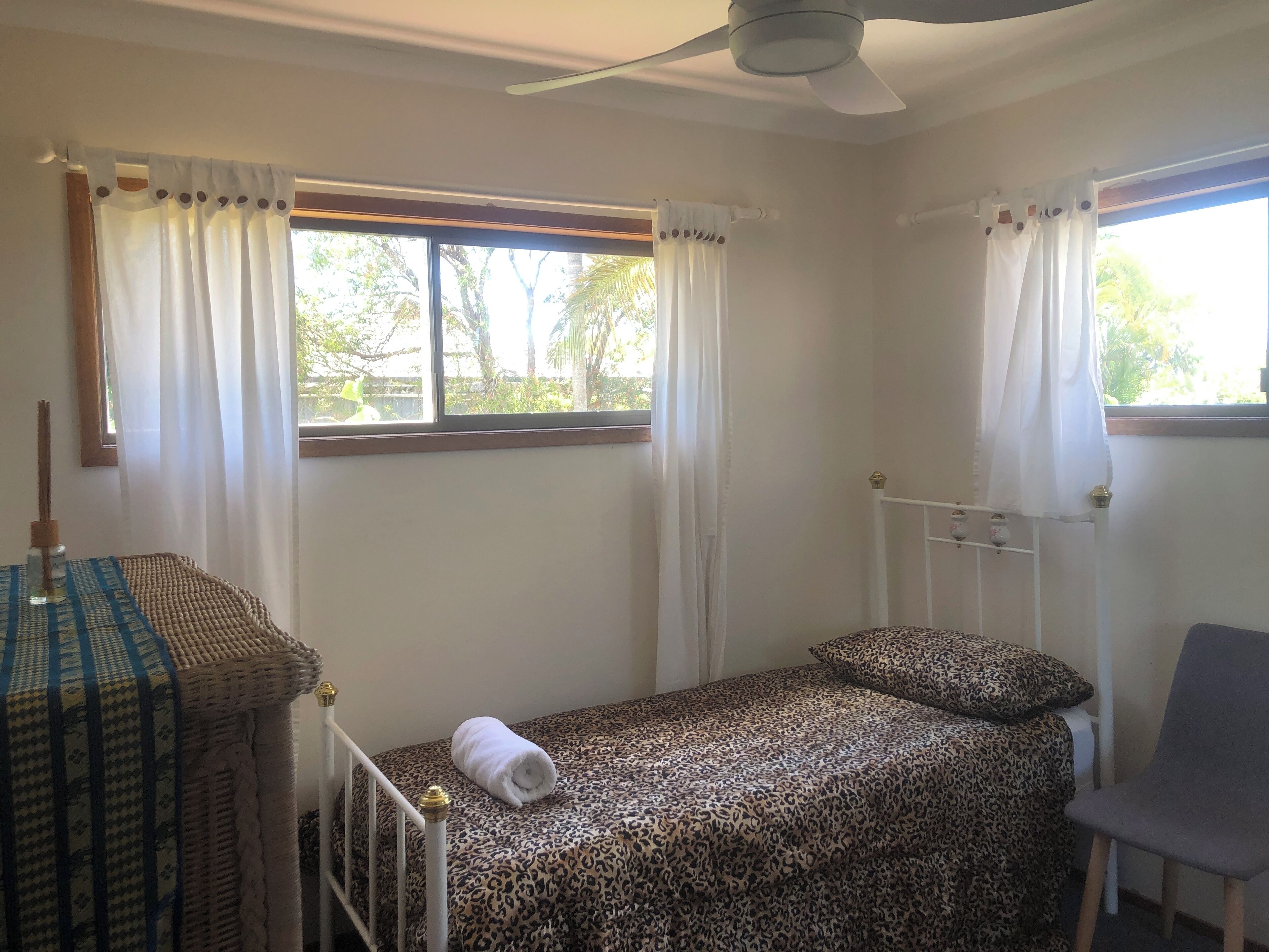 A small, single-bed room with a ceiling fan, a window with curtains, a chair, and a table with a woven tablecloth.