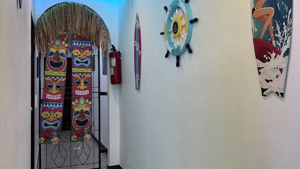 A hallway with a tiled floor, a ceiling with recessed lighting, a decorative wall with a surfboard and a steering wheel sign, and a door with colorful masks. - Broccoli candy hotels  (Karon)