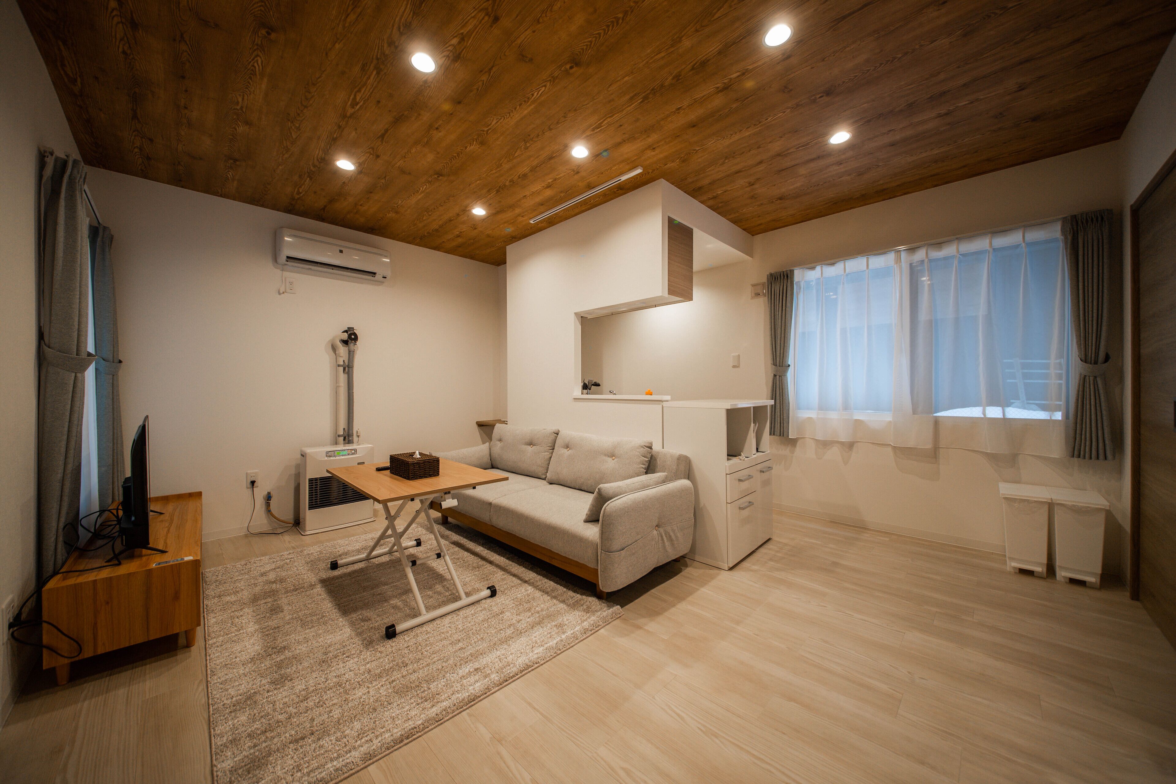 Una sala de estar moderna con techo de madera, un sofá, una mesa de centro, un televisor y una ventana con cortinas.