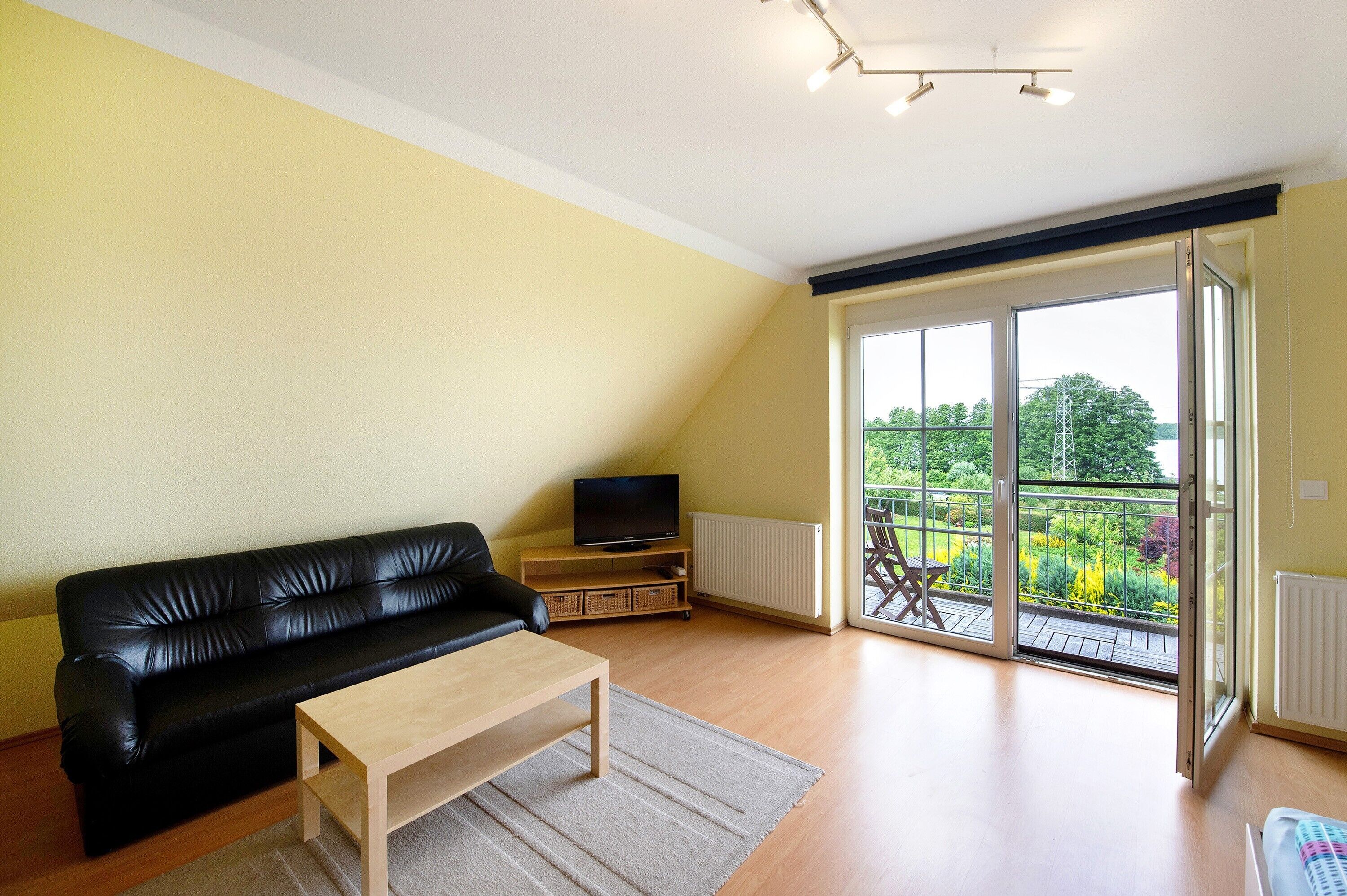 Un salón con un sofá de cuero negro, una mesa de centro de madera, un televisor sobre un mueble de madera y un ventanal corredero que da a un balcón con vistas a un jardín.