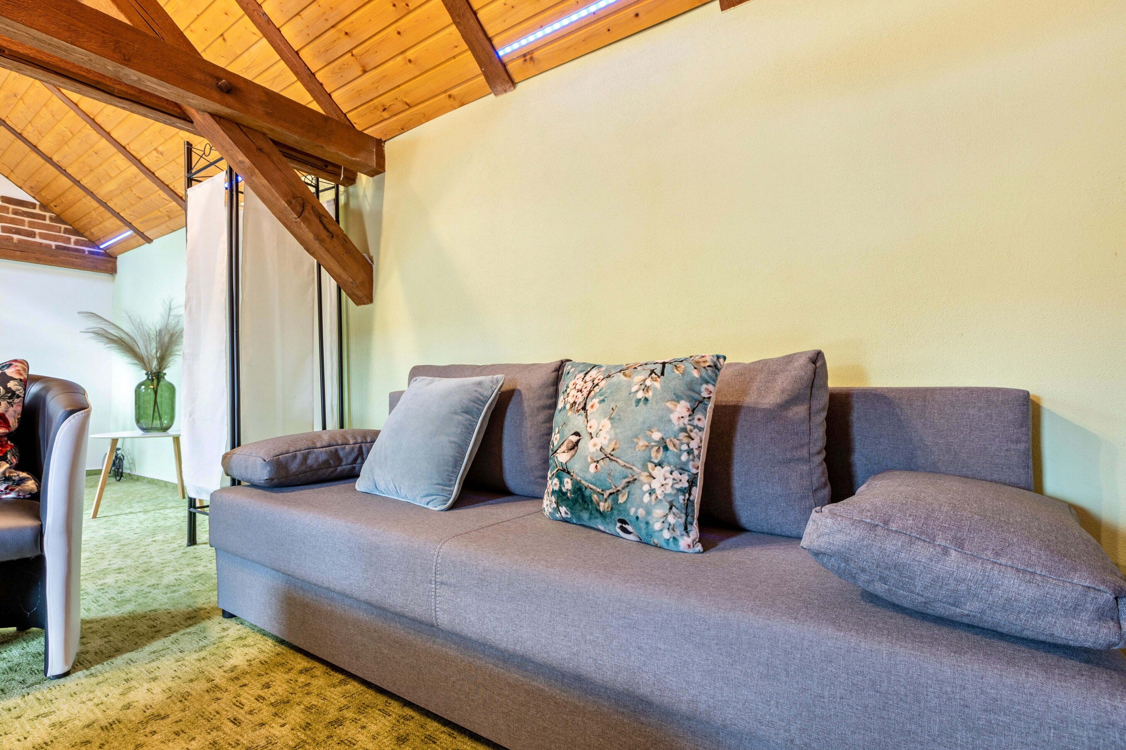 Un sofá gris con cojines florales y grises lisos, techo de madera y vistas a una habitación con una silla y un florero.