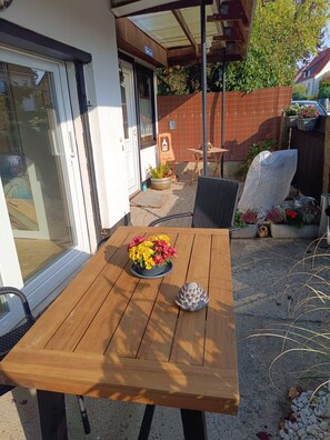 An outdoor patio with a wooden table, a potted plant with yellow and pink flowers, and a decorative stone.