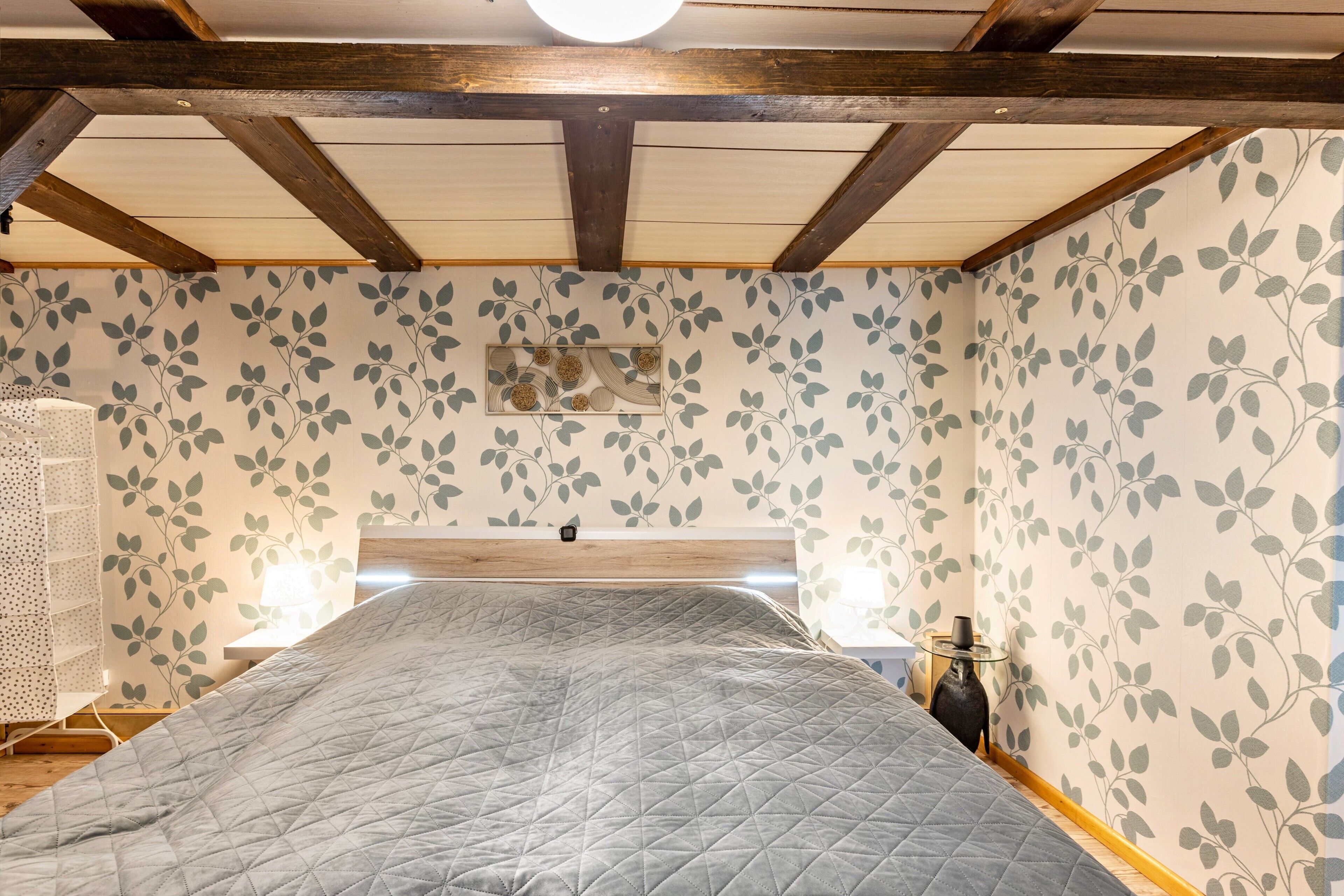 Een slaapkamer met een bed, houten balken in het plafond, een muur met bladmotieven en een nachtkastje met een lamp.