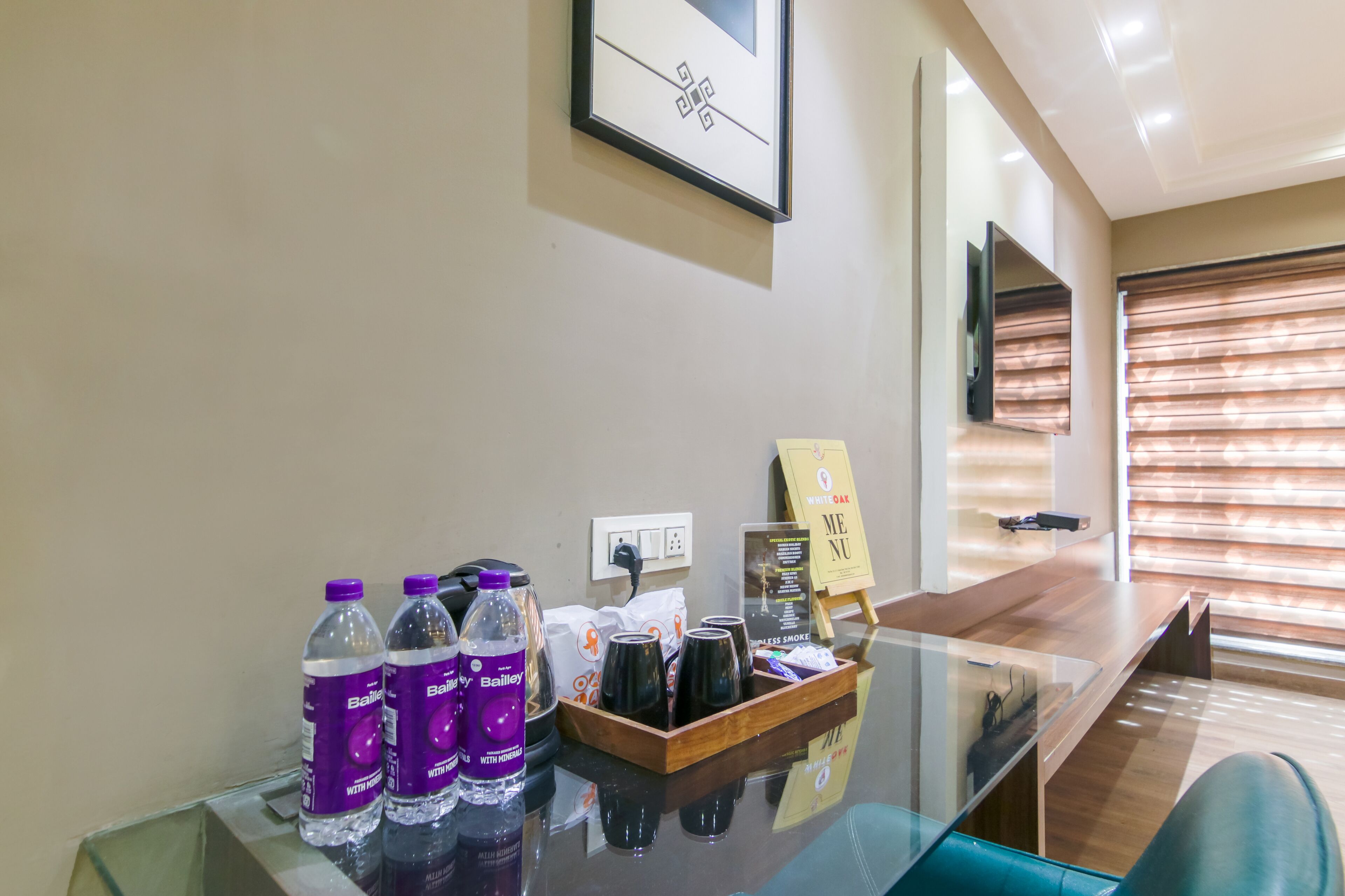 A hotel reception area with a glass table displaying water bottles, a tray with coffee cups, a menu stand, and a framed artwork on the wall.