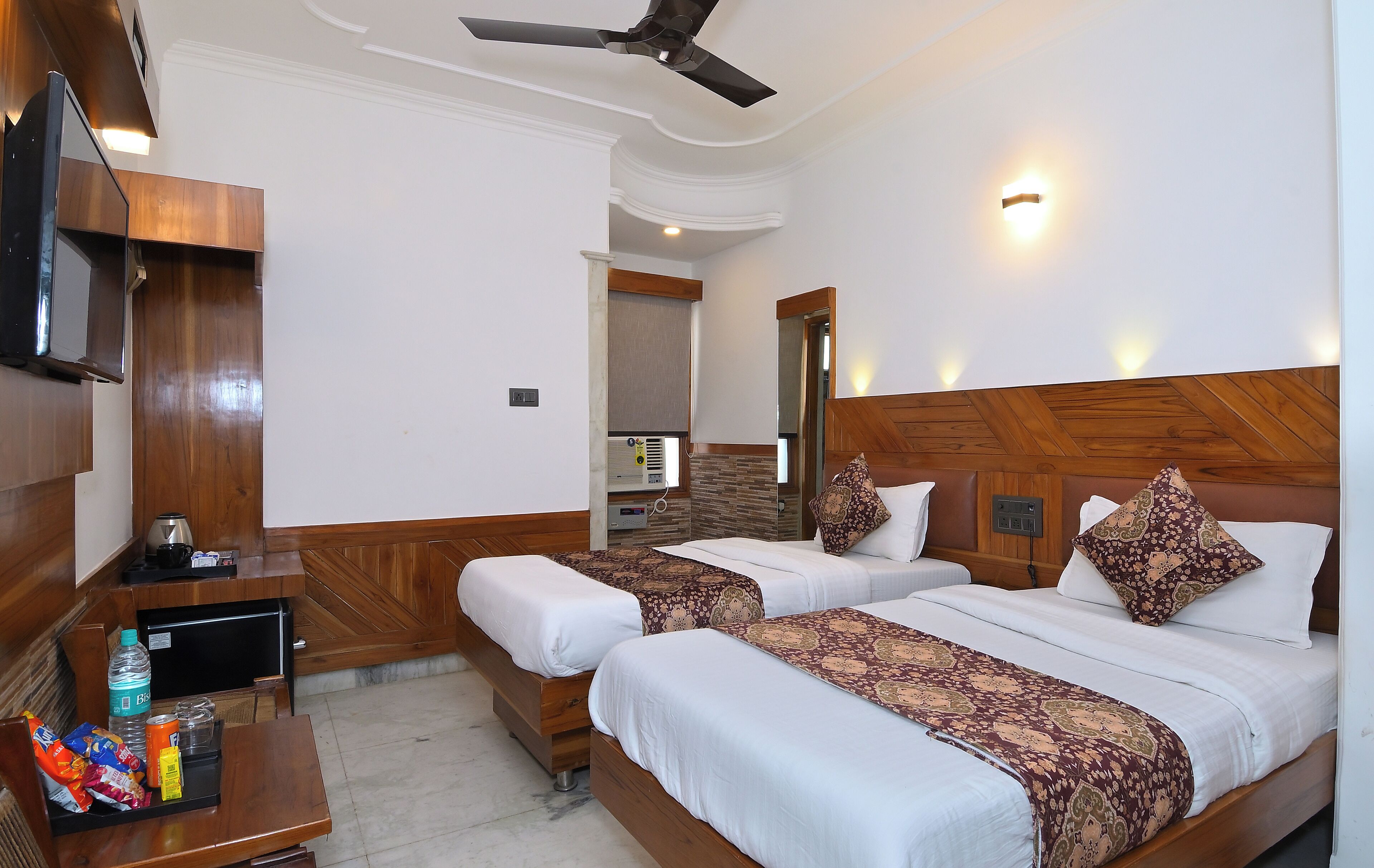 Une chambre d’hôtel avec deux lits, une télévision, un bureau avec des collations, un ventilateur de plafond et une salle de bain visible à travers une porte ouverte.
