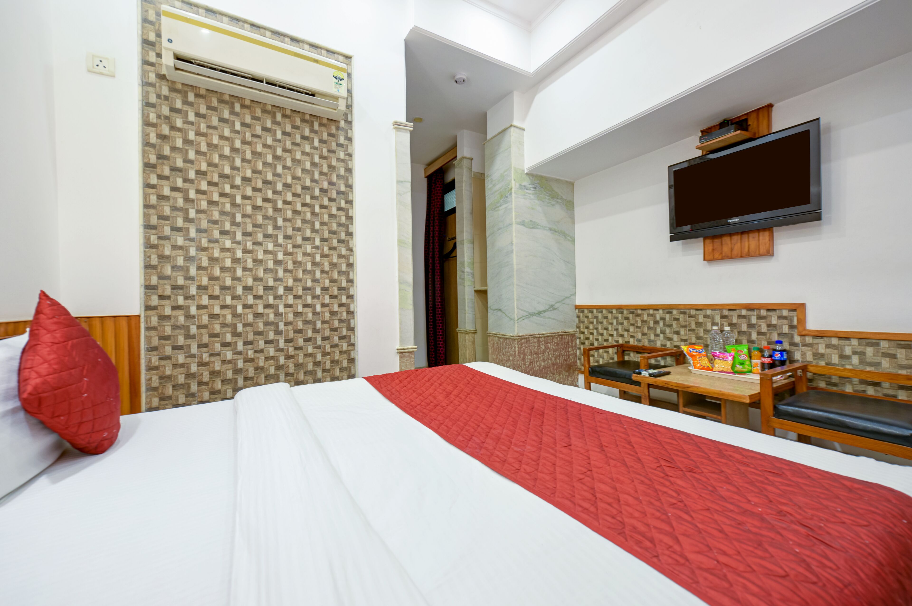 A hotel room with a bed, a TV mounted on the wall, a small table with snacks, and a red pillow.