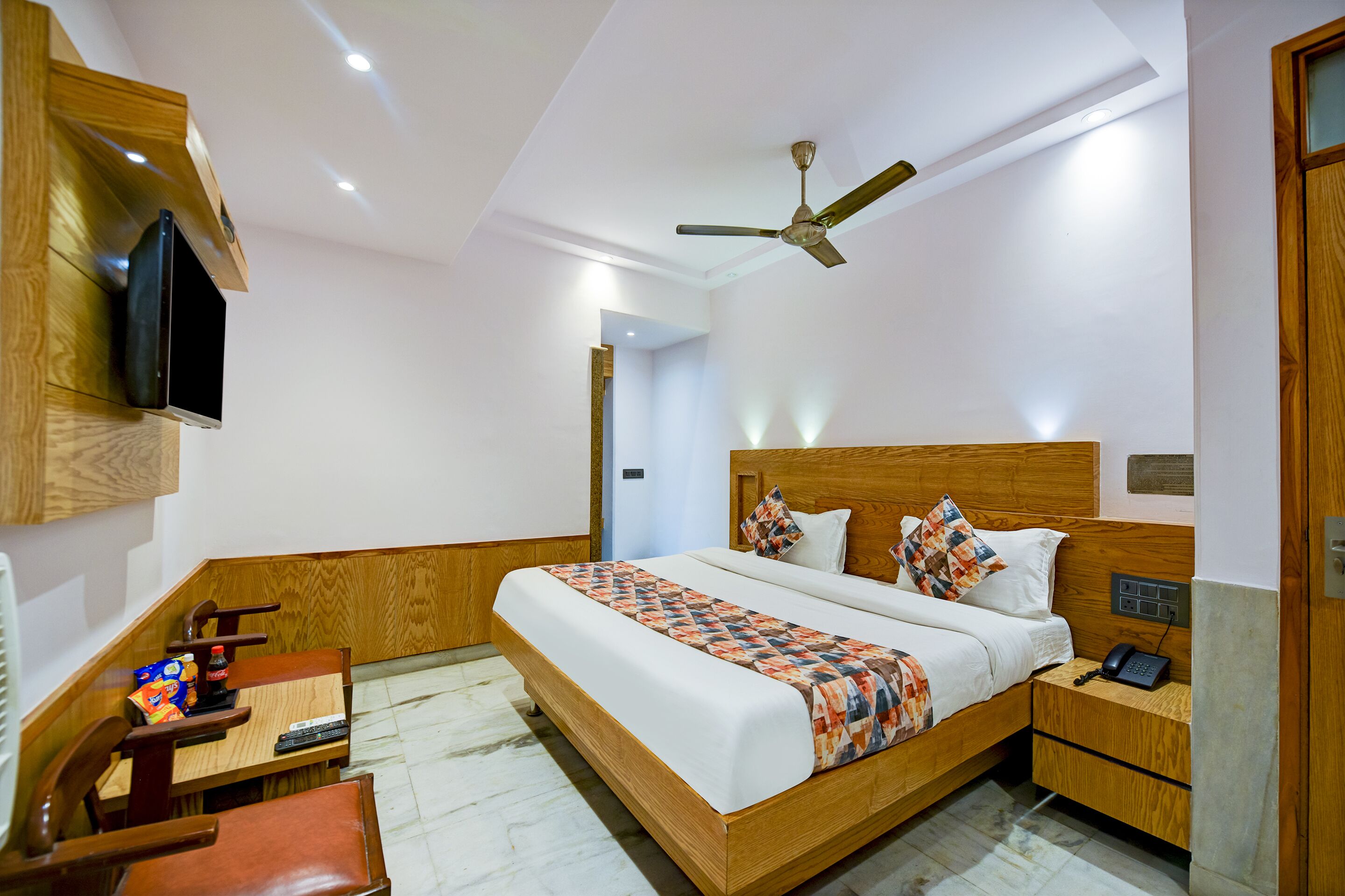 A hotel room with a wooden bed, a desk with a chair, a TV mounted on the wall, and a ceiling fan.