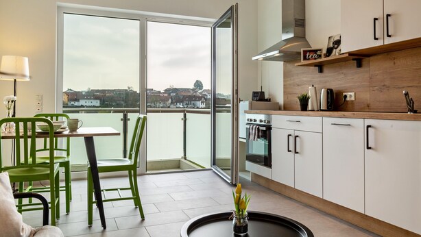A modern kitchen with a large window, a dining area with green chairs, and a view of a cityscape.