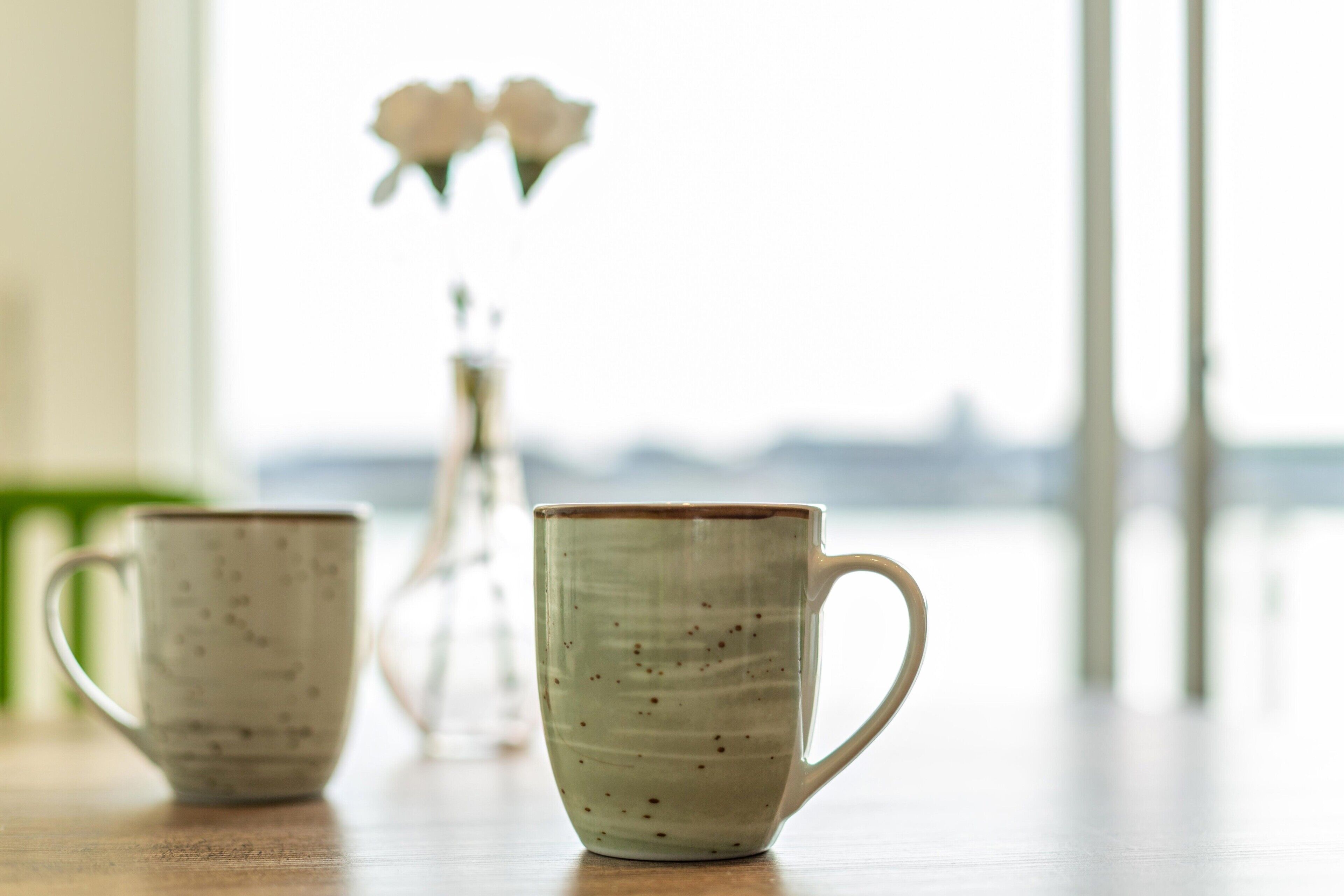 Two ceramic cups on a wooden table with a vase of flowers in the background.