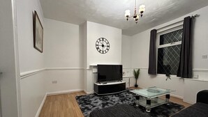 Ein Wohnzimmer mit Flachbildfernseher, einer Uhr, einer Chandelüster, einem fenster mit Vorhängen und einem Sofa.