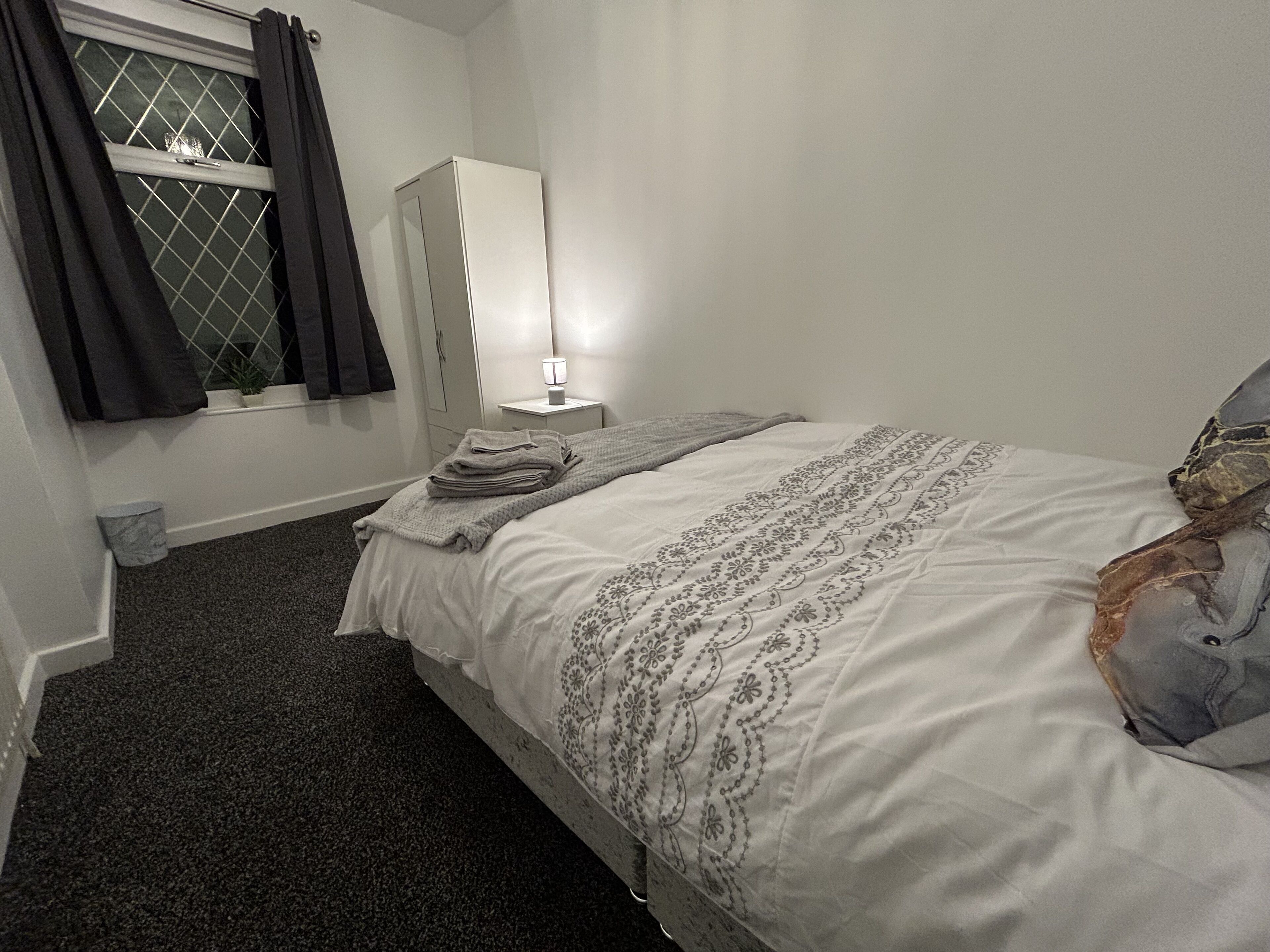 Ein Schlafzimmer mit einem Bett, einem Nachttisch mit Lampe, einem Fenster mit Vorhängen und einem kleinen Topfpflanzen.