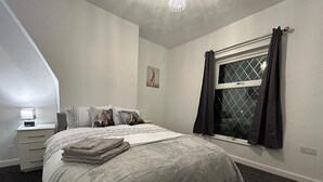 Ein Schlafzimmer mit einem großen Bett, einem Nachttisch, einem Fenster mit Vorhängen und einer kleinen Wandlampe.