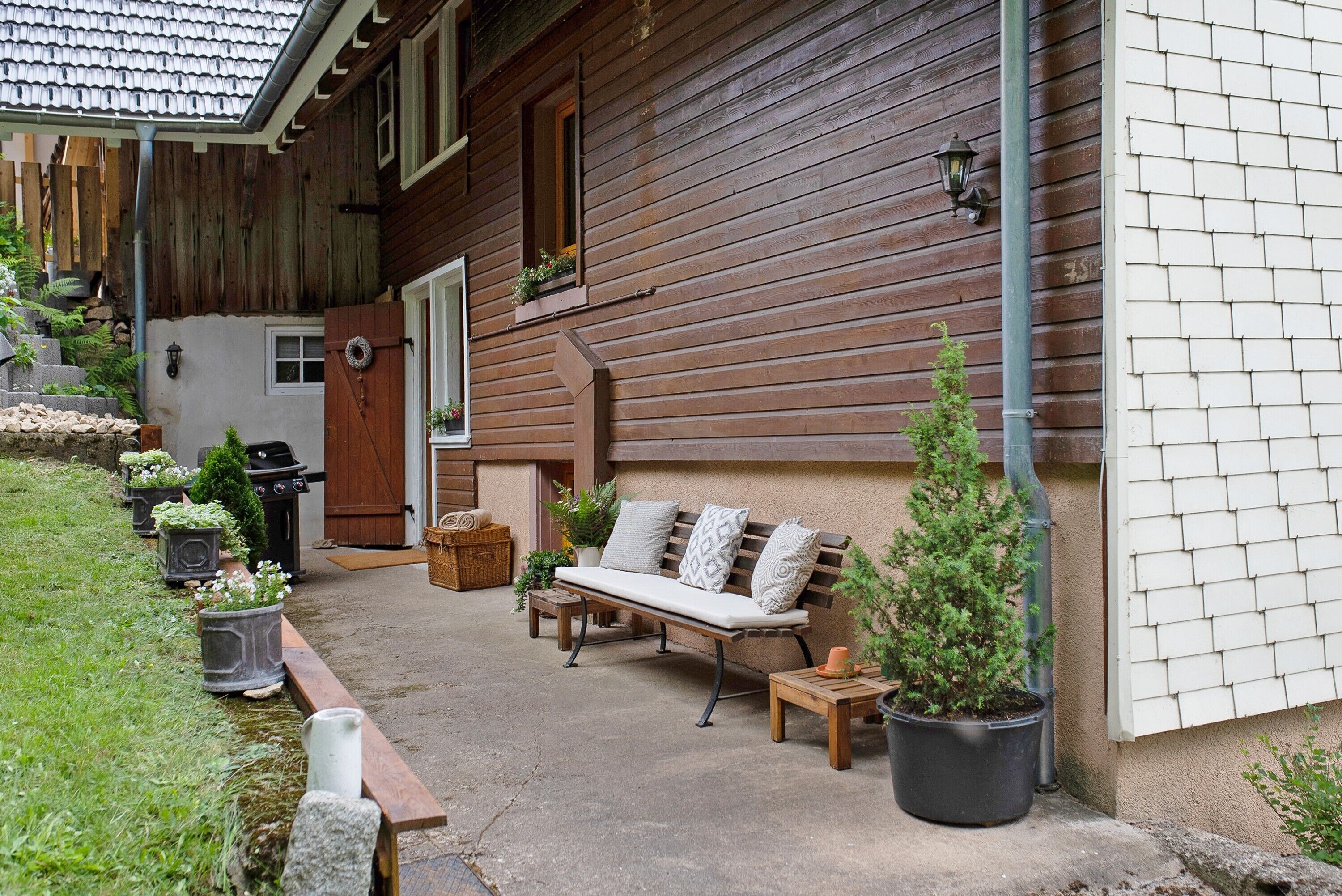 Une maison en bois avec un porche, un banc, des plantes en pot et un barbecue.