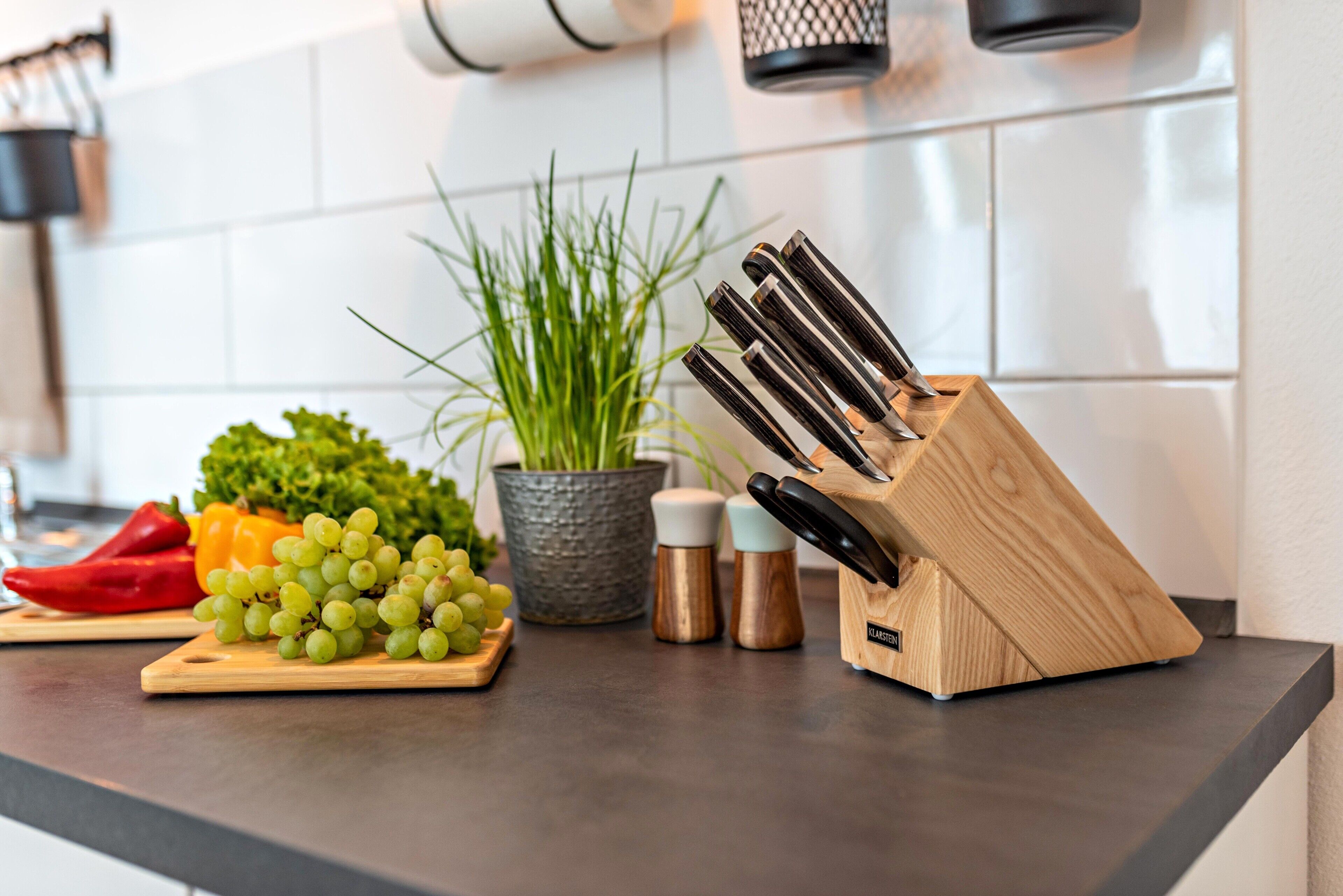A kitchen counter with a cutting board, a knife block, a potted plant, and some vegetables.