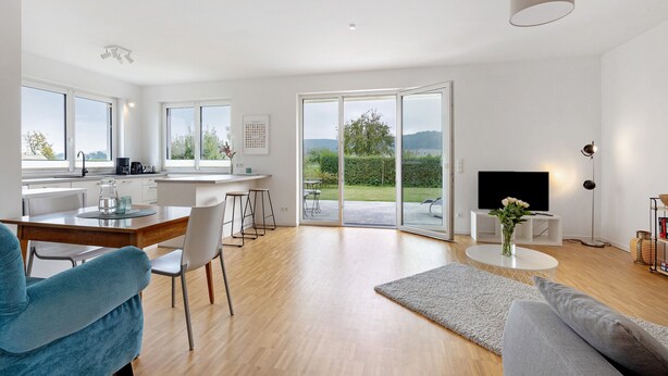 A modern living room with a sofa, a dining table, and chairs. There is a kitchen area with a coffee machine and a TV.