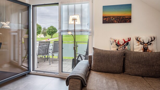 A modern living room with a sliding glass door leading to a patio, a sofa with decorative pillows, and a cityscape painting on the wall. - Apartment 'Schwarzwaldpanorama, 76qm' with Mountain View, Shared Terrace and Wi-Fi (Höchenschwand)