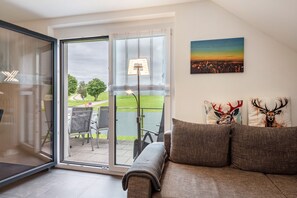 Ein modernes Wohnzimmer mit einer Schiebeglastür, die auf eine Terrasse führt, einem Sofa mit Zierkissen sowie einem Stadtbild an der Wand.